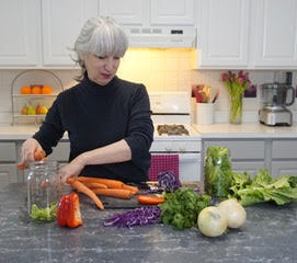 Woman in a kitchen, in front of her is carrots, cabbage, lettuce and a empty mason jar